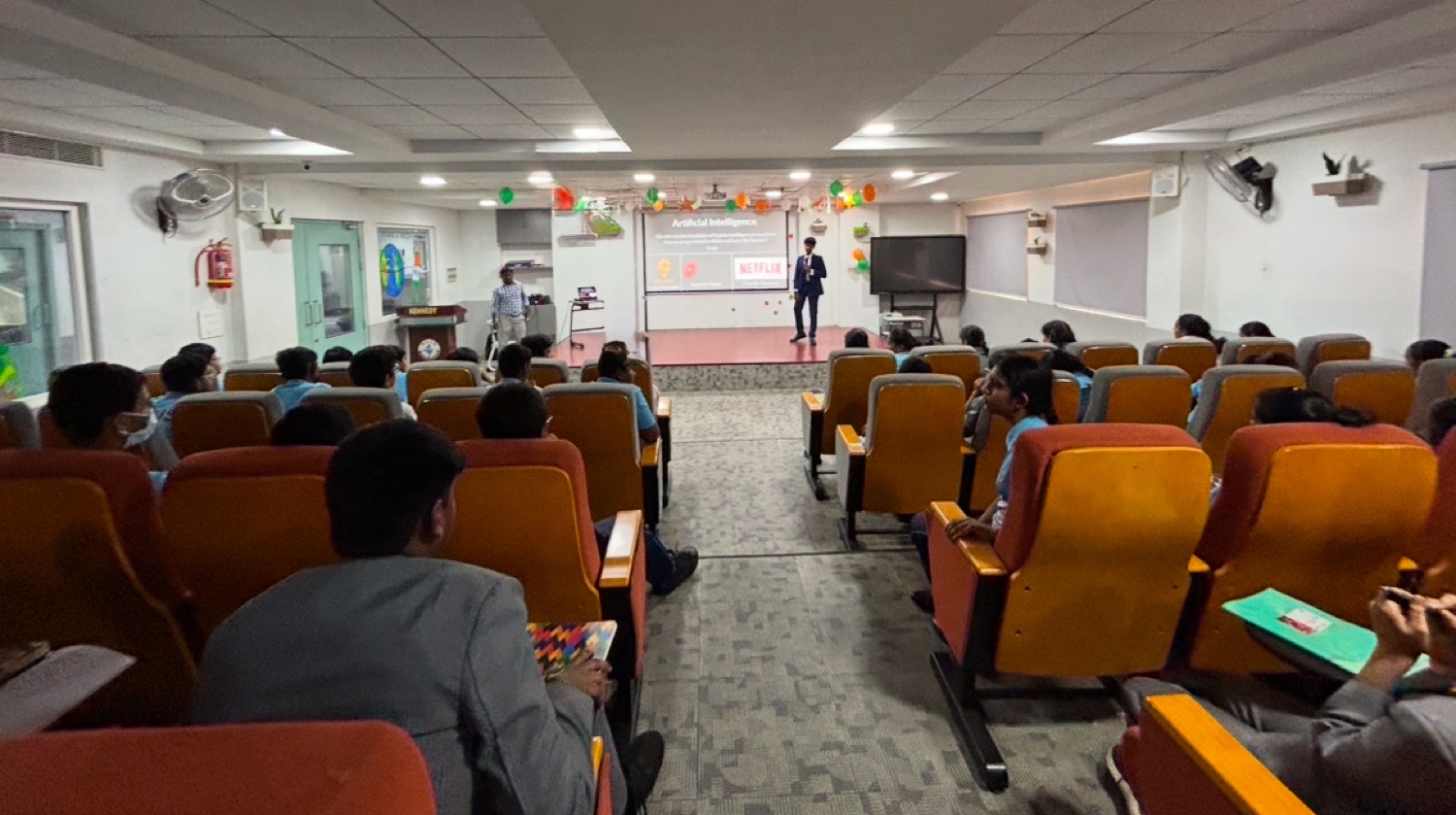 Wide view of an auditorium, rows of seated students facing the stage with Shashank presenting